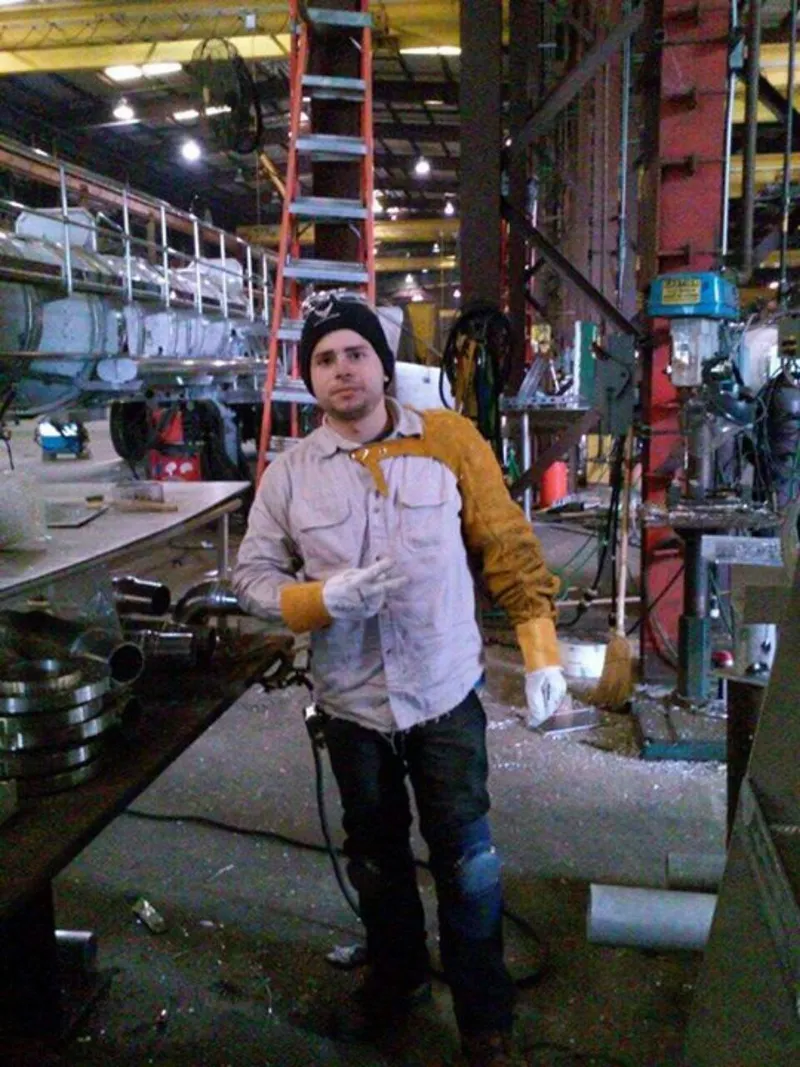 David Cruz in the Dragon Products plant in Texas, in a beanie, work shirt and welding gloves with a leather sleeve over one shoulder, a line of steel tanks under fabrication behind